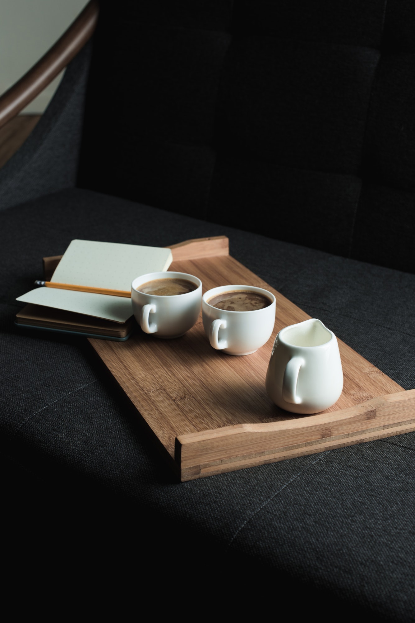 two-cups-of-coffee-jug-of-milk-and-notebook-with-pencil-on-wooden-tray-on-sofa